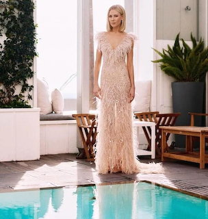 Woman in a pale feathered gown standing beside a turquoise pool in a bright courtyard