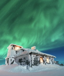 Snow-covered house beneath green northern lights in a starry winter sky