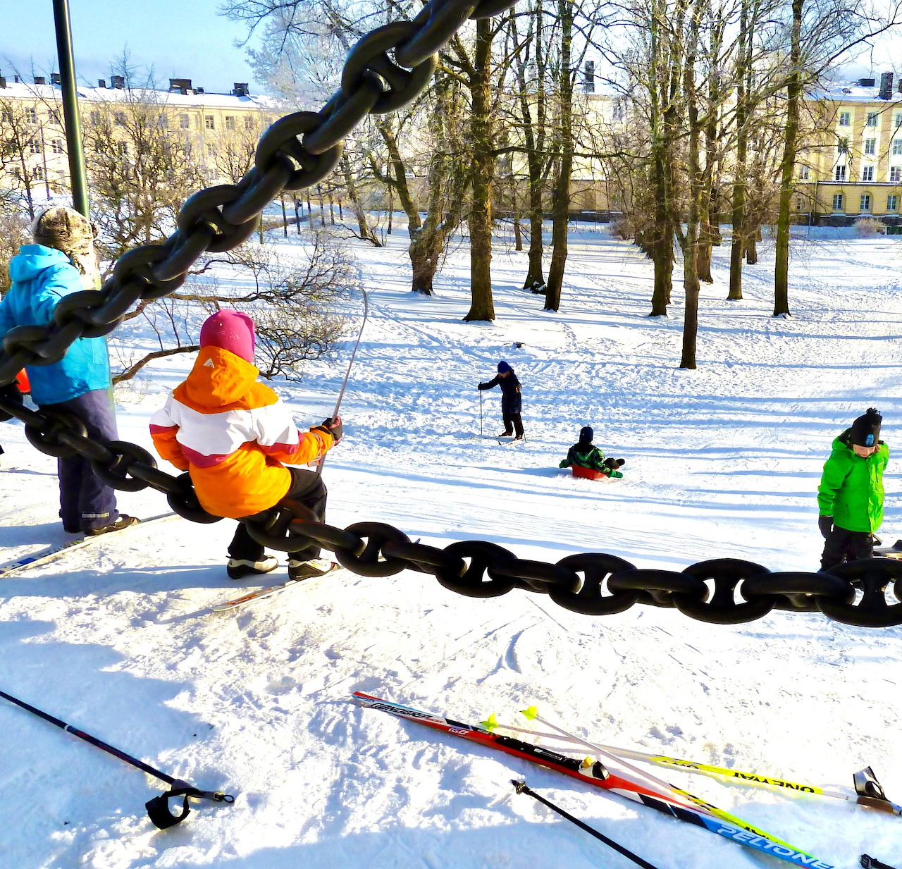 Children playing in a snowy park near a giant chain swing with bare trees and skis