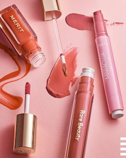 Assorted pink and coral lip glosses and lipsticks arranged on a pink background with color swatches