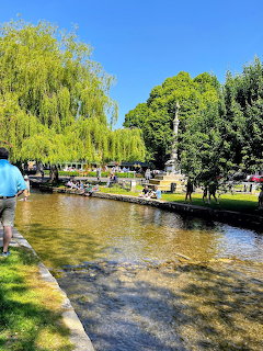 Shallow river with stone banks flowing through green park with willow trees and visitors on sunny day