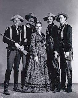 Five actors in Western cavalry uniforms and 19th century dress pose together in a vintage black and white studio photograph