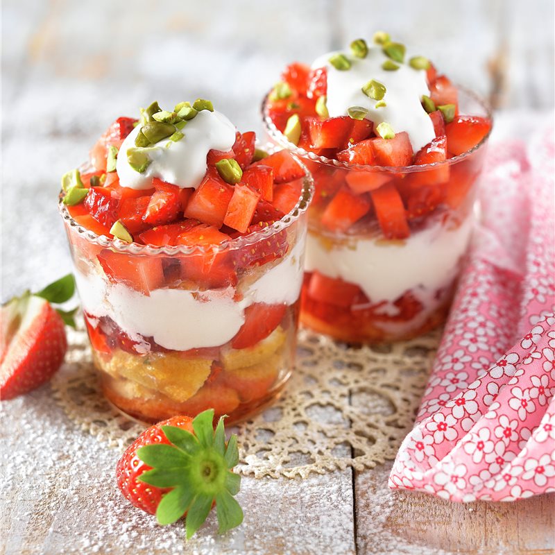 Two layered strawberry dessert cups with cream, cake, and pistachios on a rustic table