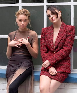Two women in formal attire posing together against a white paneled backdrop