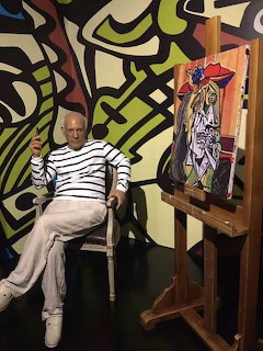 Wax figure of Pablo Picasso in striped shirt sitting next to an easel with a cubist painting