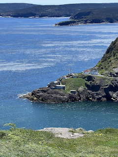 Rocky coastal headland with small buildings overlooking blue ocean water and green hills
