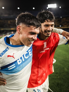 Two young soccer players in white and red jerseys celebrating together on a pitch at night