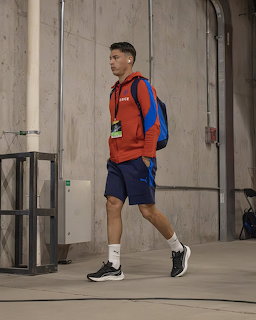 Athlete in orange jacket and blue shorts walking with backpack in concrete indoor space