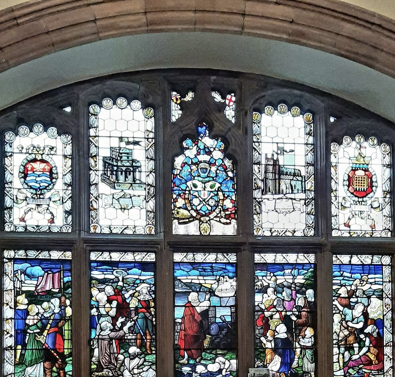 Arched stained glass church window with colorful figure panels and heraldic designs