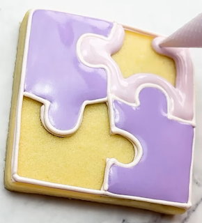 Square sugar cookie decorated with purple and yellow jigsaw puzzle piece icing design being piped with pink icing