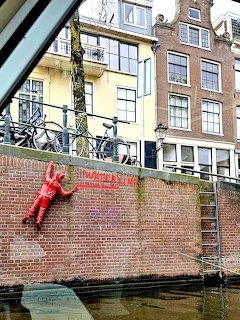 Red climbing sculpture on a brick canal wall with bicycles and Amsterdam buildings above