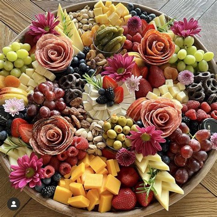 Circular platter with grapes, berries, cheese cubes, salami roses, and pink flowers arranged decoratively
