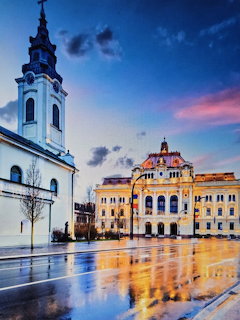 Twilight view of Oradea town square with baroque church and ornate buildings reflected in wet plaza