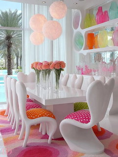 White modern dining room with sculptural chairs, colorful cushions, pastel rug, and rainbow glassware on shelves