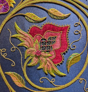 Detailed embroidery showing a stylized flower with pink petals and gold accents on blue background with golden swirls
