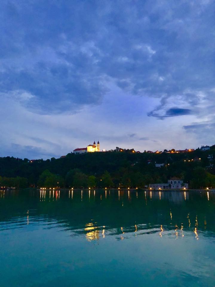 Illuminated monastery on hilltop at dusk with water reflection and blue sky