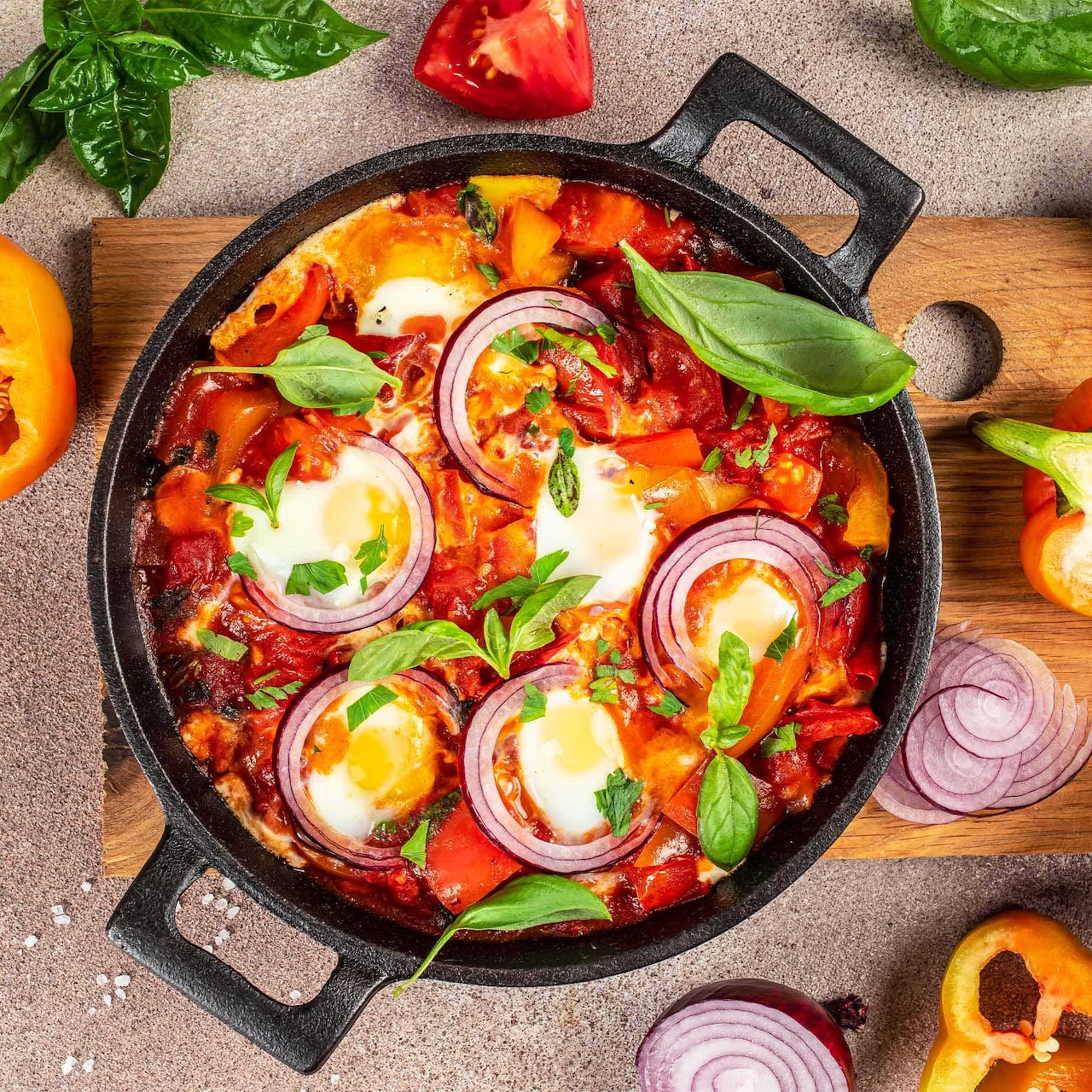 Shakshuka in black cast iron pan with eggs, peppers, onions and basil on wooden board