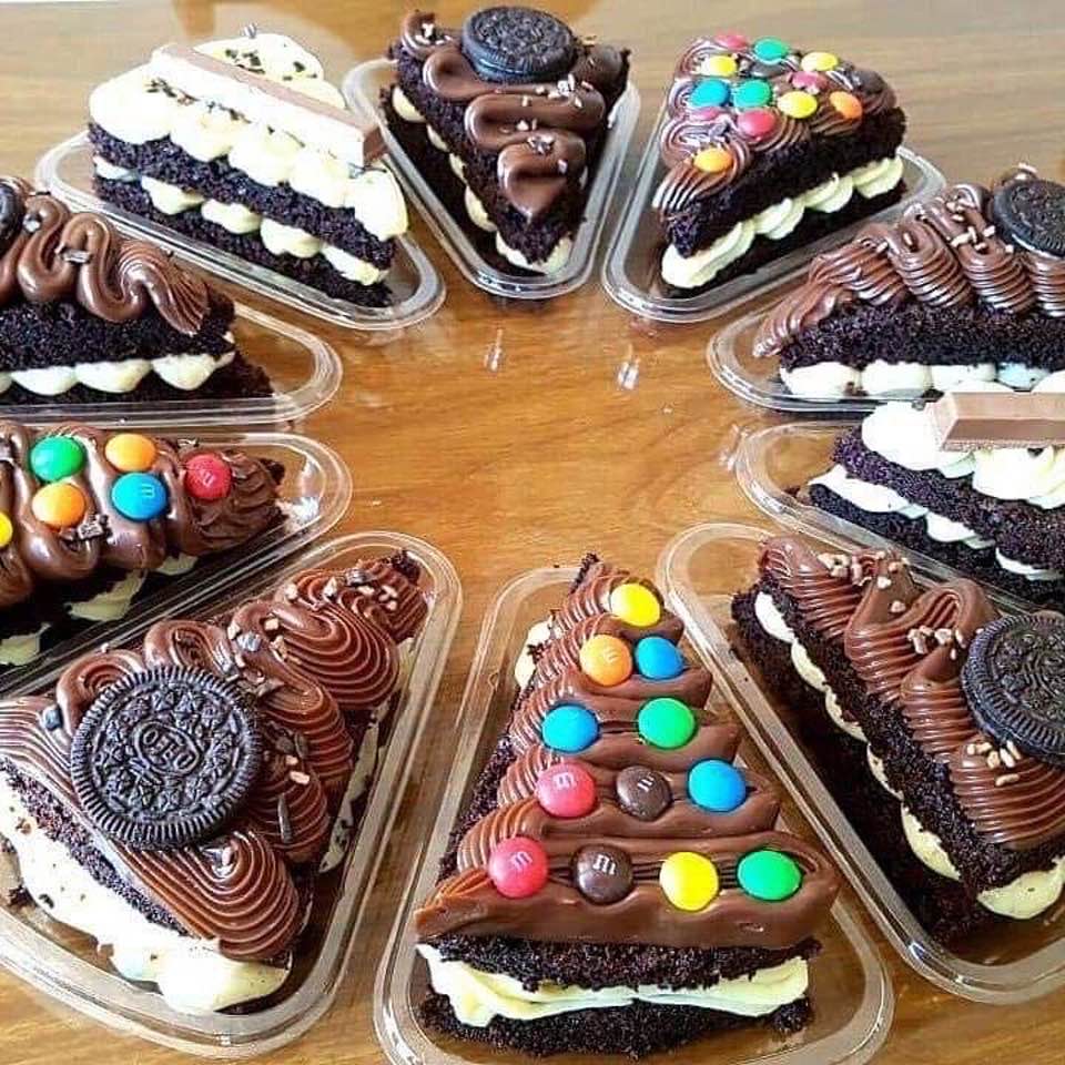 Various chocolate cake slices with frosting, Oreos, and candy toppings in plastic containers on wood surface