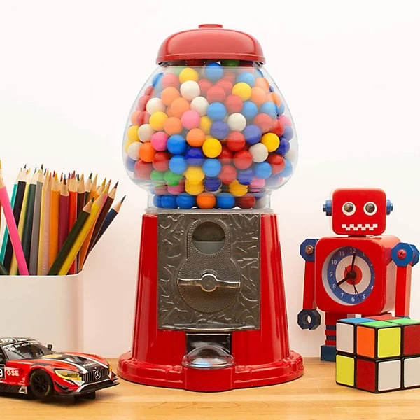 Red vintage gumball machine with colorful candy balls on desk surrounded by toys and pencils