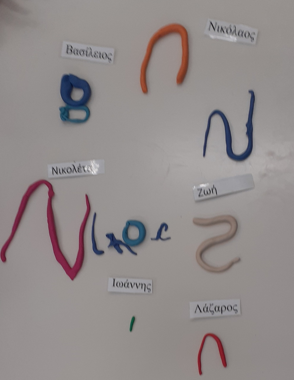 Colorful clay letters forming initials of Greek names on a white surface with name labels