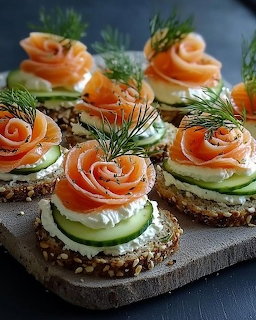 Seeded crackers topped with cream cheese, cucumber, smoked salmon roses, and dill sprigs on a wooden board