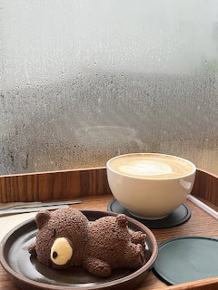 Chocolate teddy bear dessert on a plate beside a latte by a rainy window