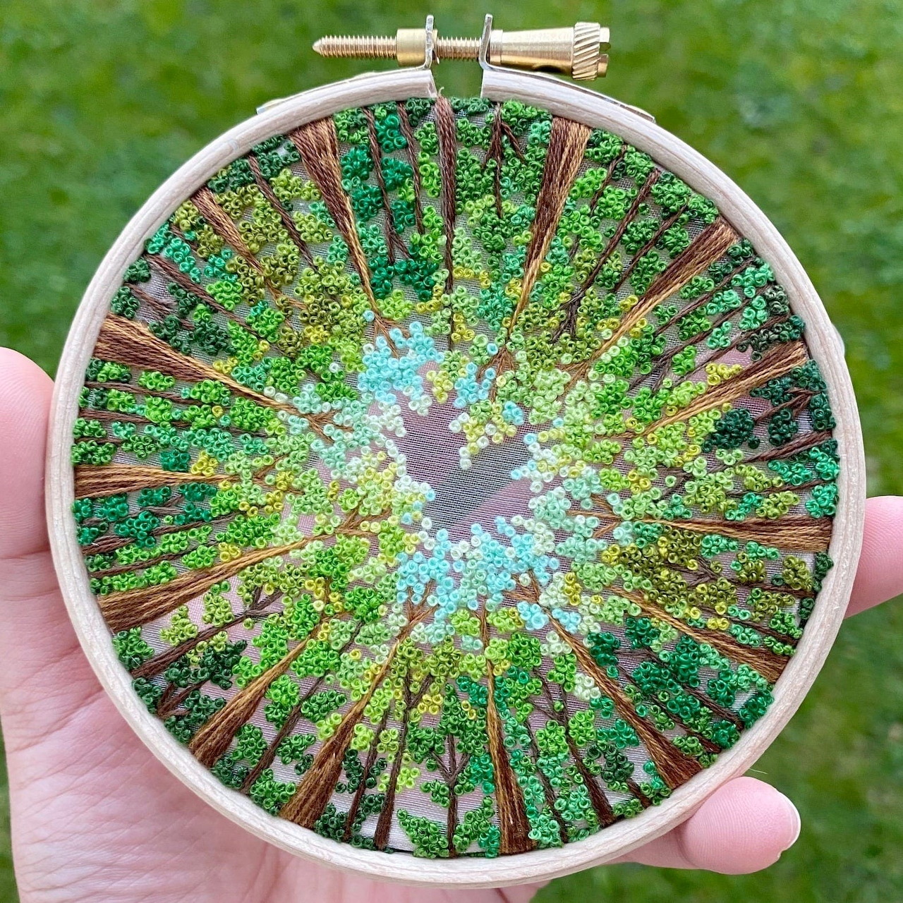 Hand-held embroidery hoop with French knot forest design in green and blue threads against blurred garden background