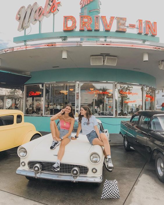 Two women sitting on a white vintage car in front of a retro drive-in with classic cars nearby.