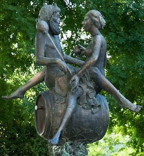 Bronze statue of two figures sitting on a large urn beneath green trees