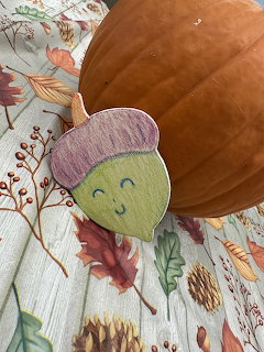 Smiling acorn craft next to a large pumpkin on autumn leaf-patterned fabric