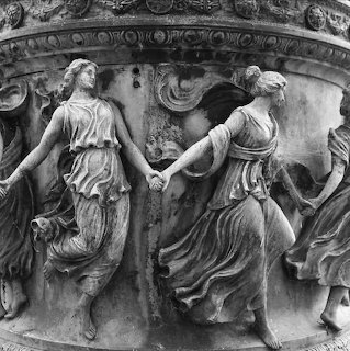 Black and white stone relief of draped women dancing in an ornate circular frieze