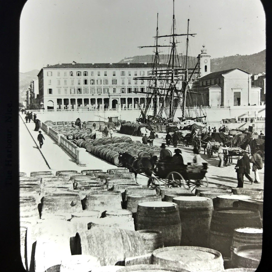 Black and white photo of 1889 Nice harbor with sailing ship, workers, barrels, and buildings along the waterfront