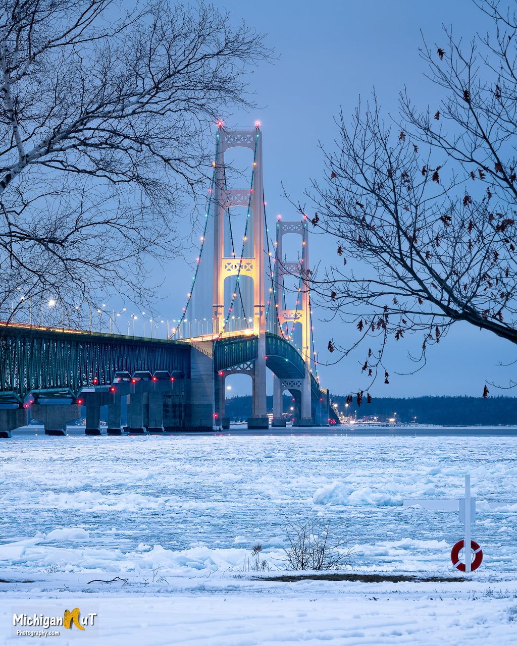 Mackinac Bridge at dusk over icy water, framed by leafless trees