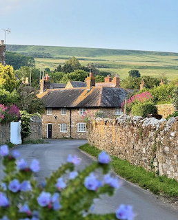 Stone cottage by a village road with flowers, stone walls, and green hills behind