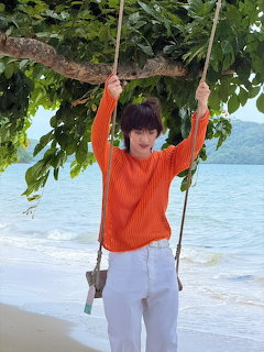 Person in an orange sweater on a rope swing under a tree by the beach