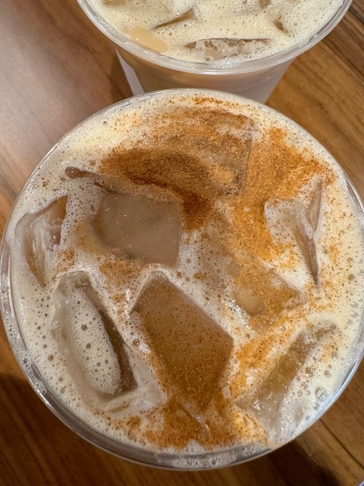 Top view of two iced coffee drinks with foam and cinnamon in clear plastic cups on a wooden table