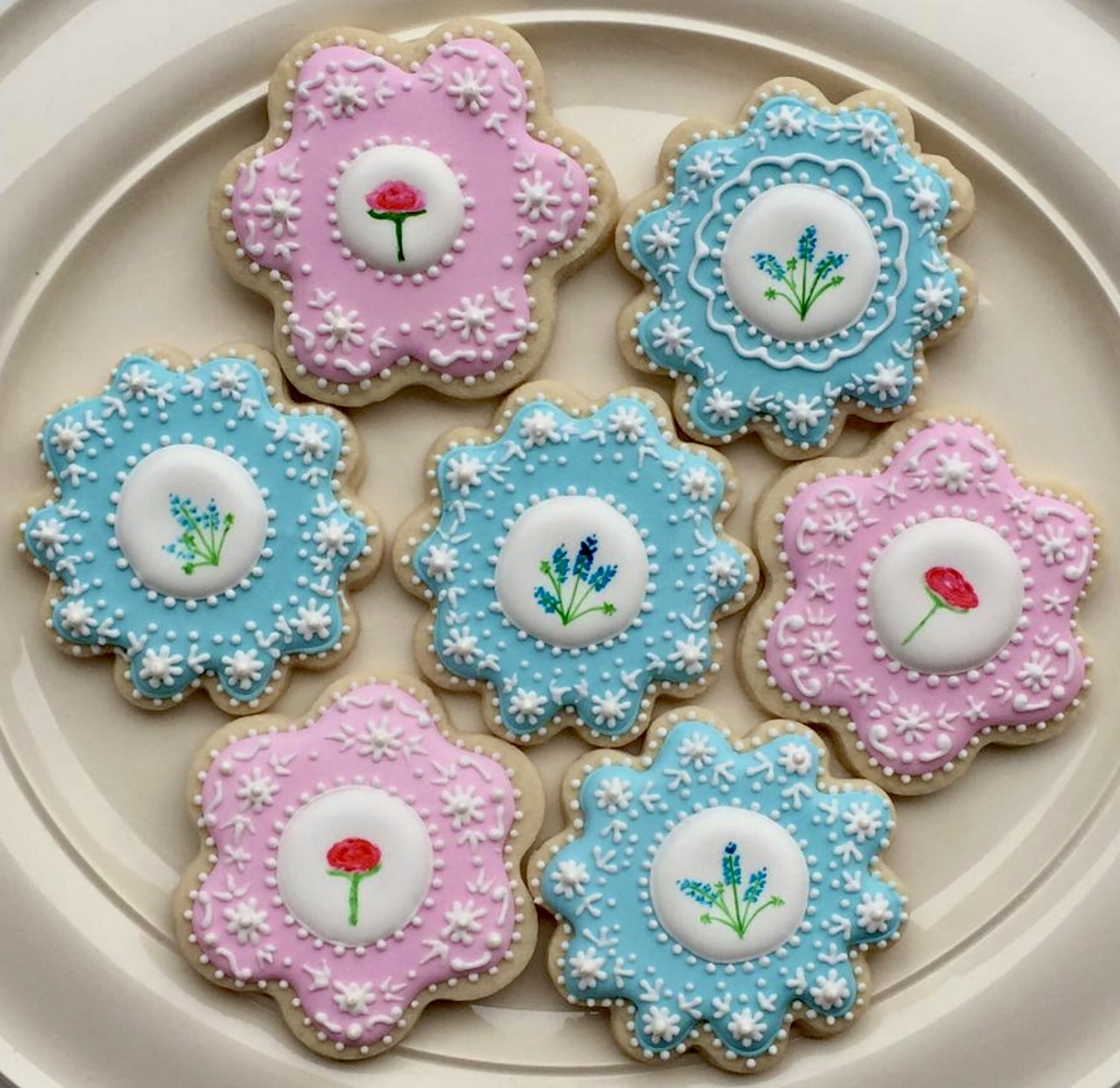 Six pastel flower-shaped decorated cookies on a plate with floral icing designs