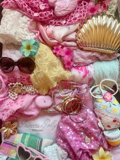 Flatlay of pink and pastel summer accessories including sunglasses, pearls, lace, flowers, and a gold fan