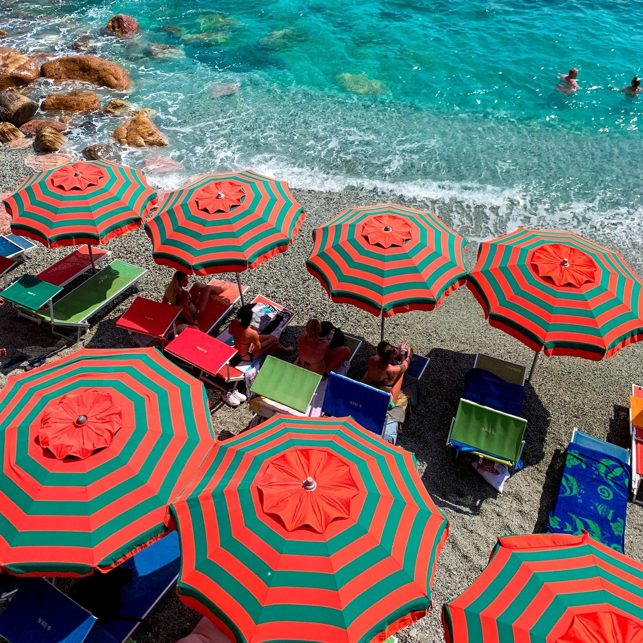 Overhead view of red and green striped beach umbrellas on rocky Italian coastline with crystal clear turquoise water