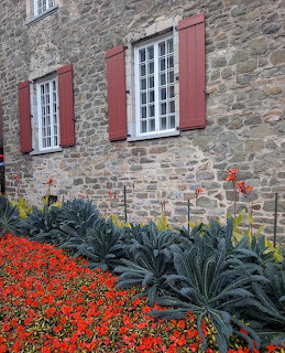 Stone building with red shutters and white windows above a garden of red flowers and ornamental kale