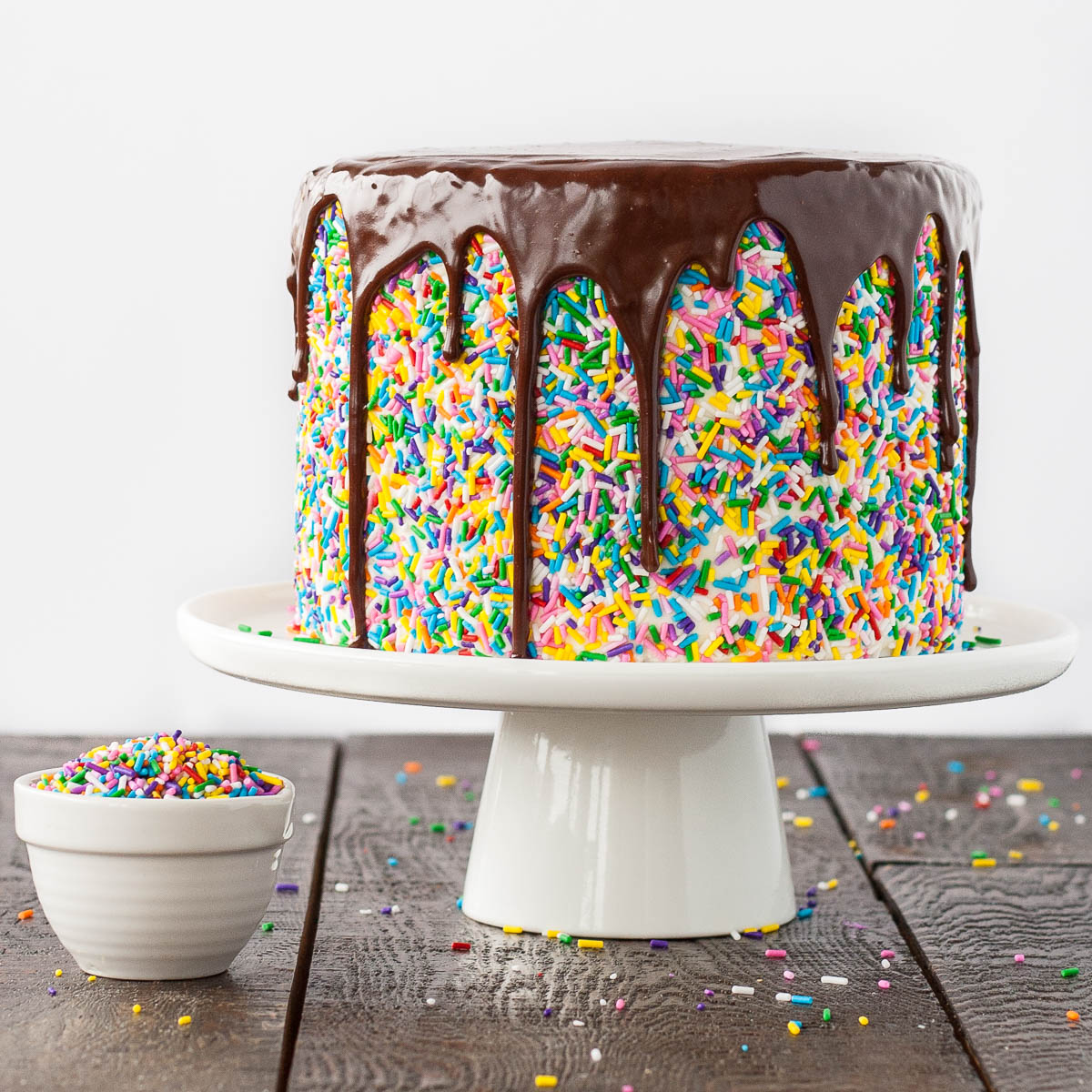 Funfetti cake with chocolate drip on white cake stand with bowl of sprinkles
