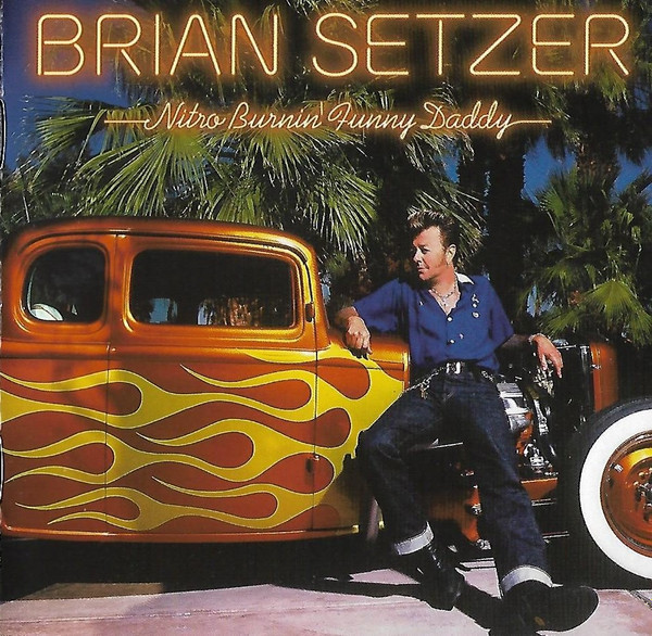 Brian Setzer sitting beside a vintage flame-painted hot rod under palm trees at night