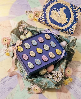 Blue gift box containing pastel-colored chocolate eggs arranged in rows, surrounded by flowers and ribbon