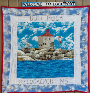 Quilted wall hanging of Gull Rock with a lighthouse-like building, rocks, blue water, and cloudy sky