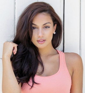 Young woman with long dark hair in a pink tank top posing against white wooden wall