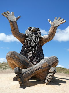 Wooden sculpture of a seated bearded figure with raised arms under a blue sky