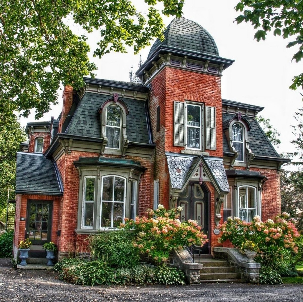 Victorian red brick house with turret, mansard roof, white trim, and flower garden in front
