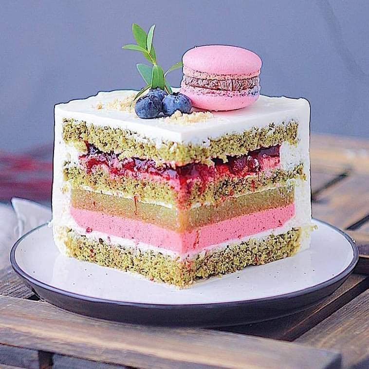 Square layered cake slice with pink macaron, blueberries, and mint garnish on white plate