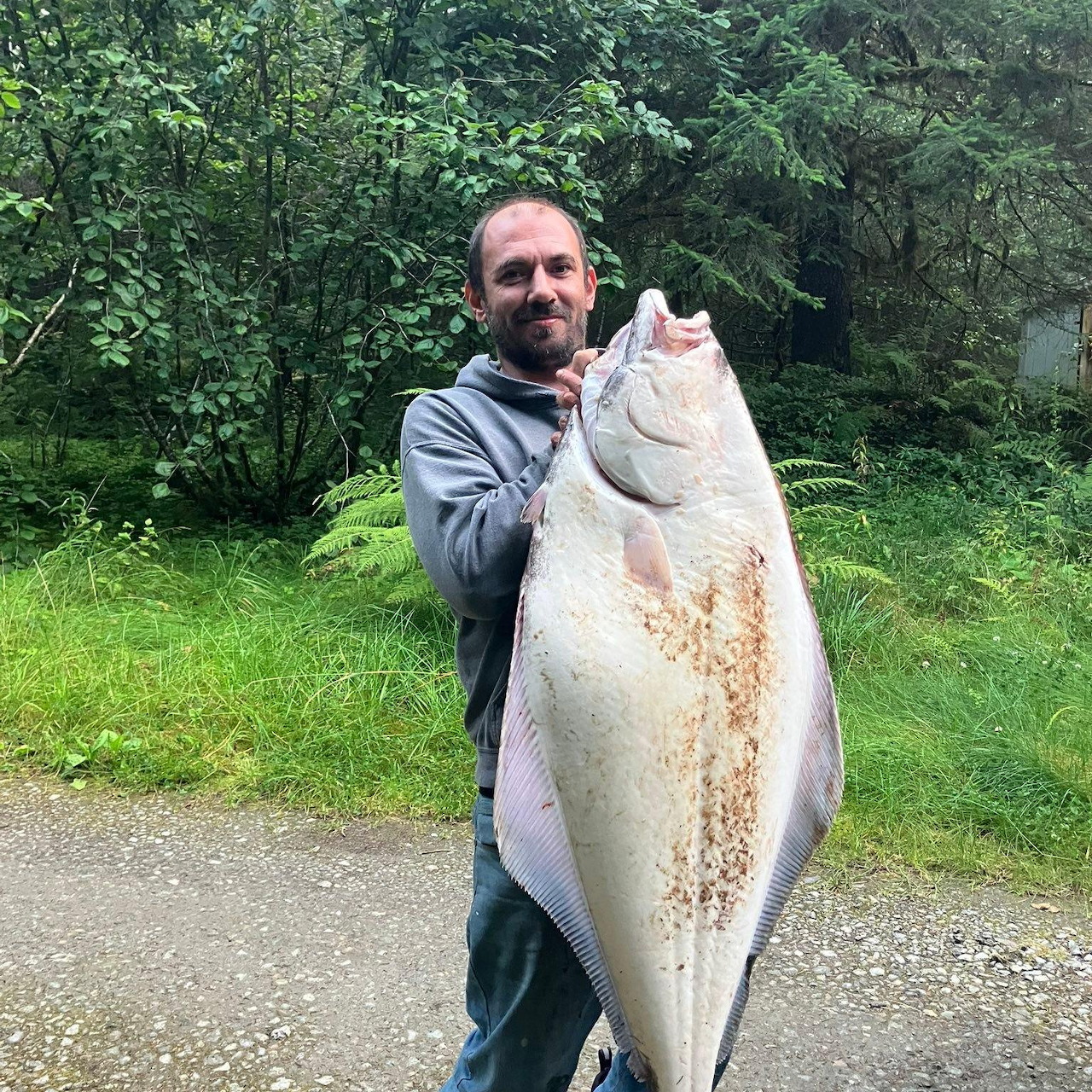 Man holding a large white Pacific halibut fish outdoors with trees in background