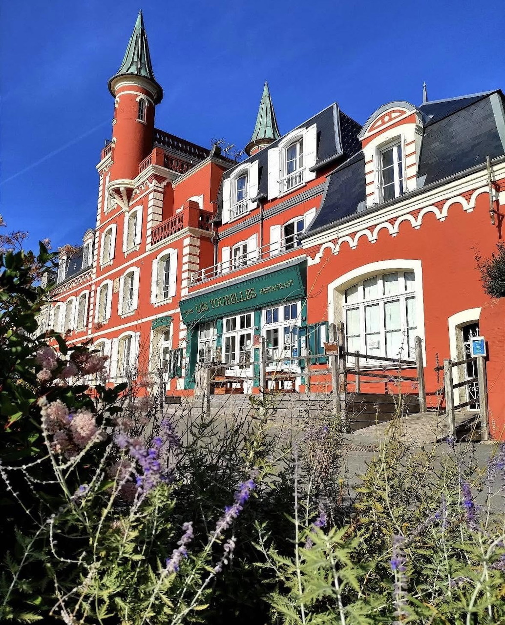 Red historic house with small turrets, white-trimmed windows, and garden plants in front